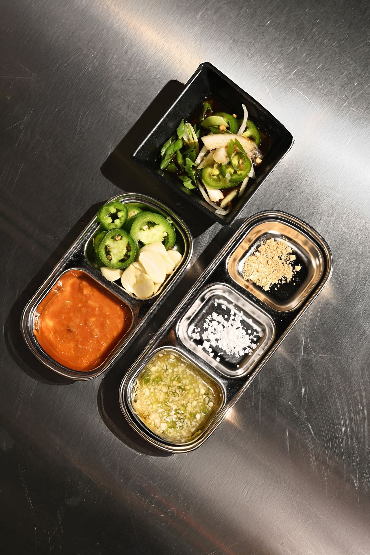 Various spices and sauces in small metal trays on a stainless steel surface.